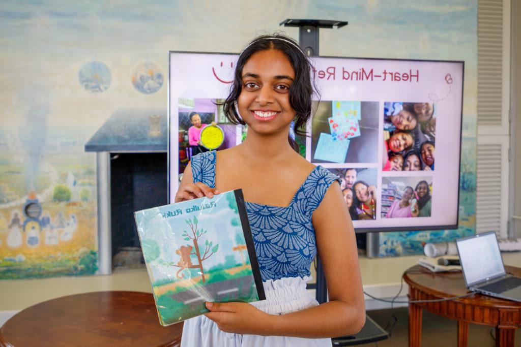 Pratya Poosala holds up her children’s book titled Rajulko Rukh. The book cover is a digital illustration of a brown monkey sitting on a tree by a street. Poosala has dark wavy hair pulled back and wears a blue top and white pants.
